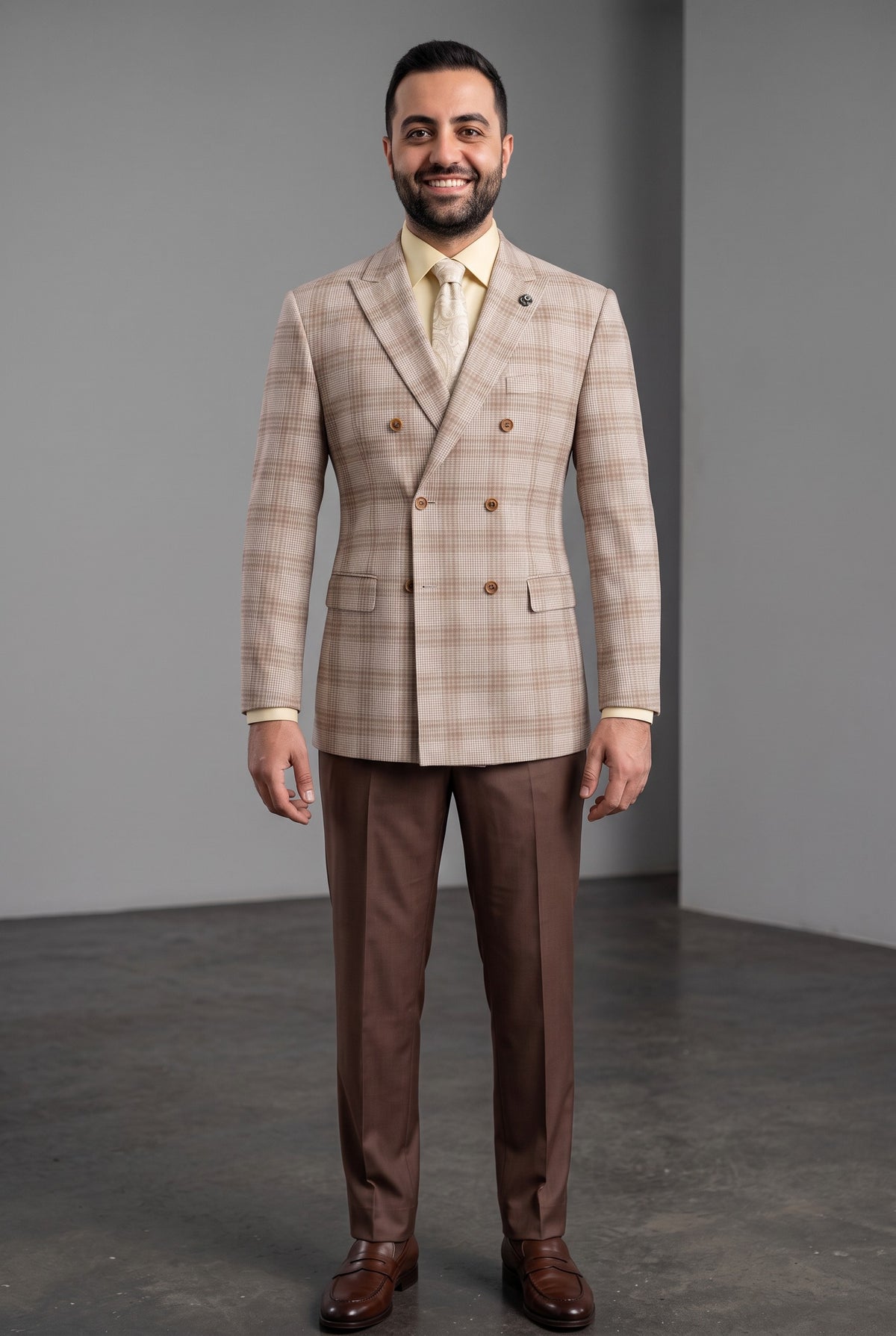 A man modeling a beige and brown plaid double-breasted suit jacket with matching brown trousers, a light yellow tie, and brown leather dress shoes, standing against a gray studio background.