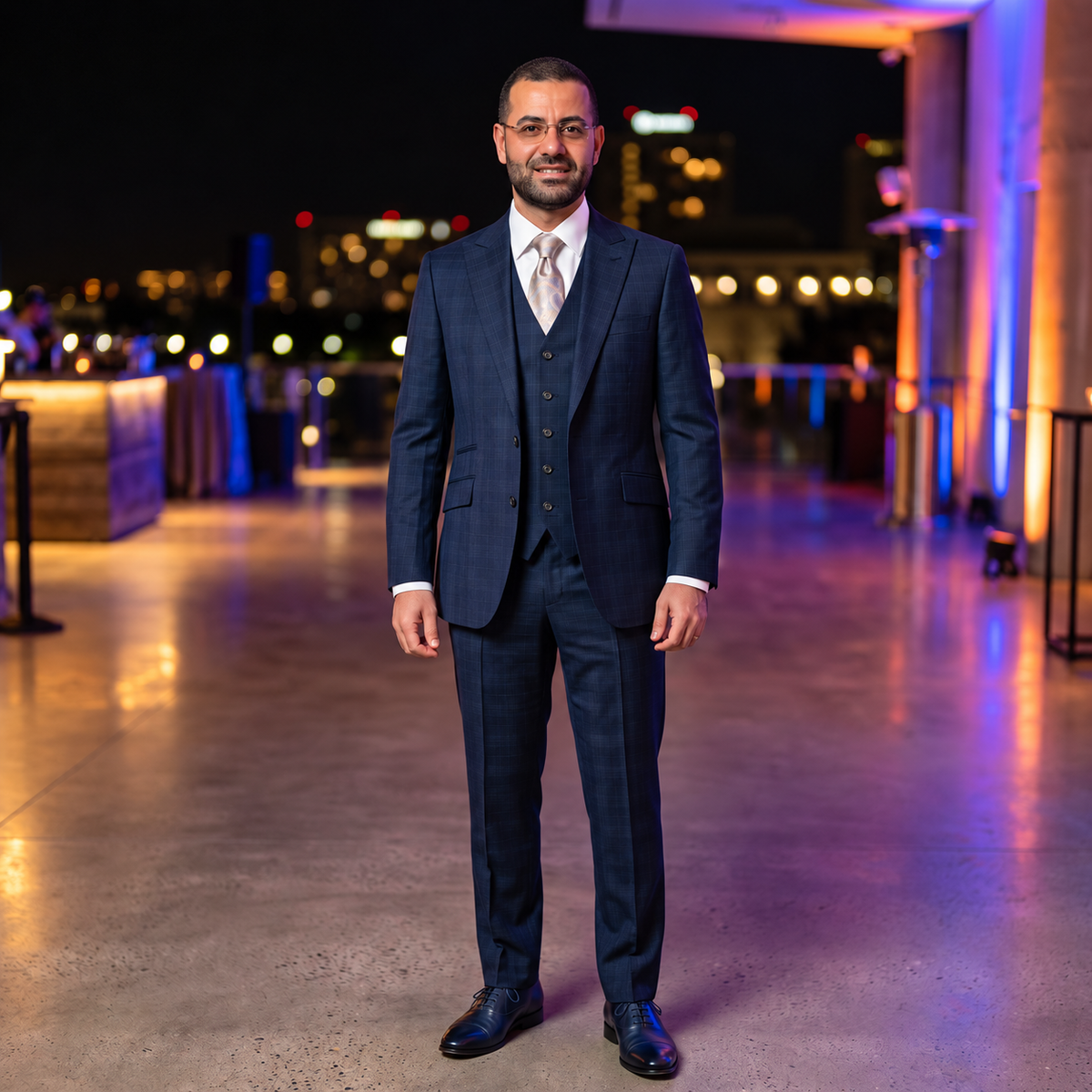 A man wearing a dark navy blue three-piece suit with a white dress shirt and a light-colored tie, standing on a rooftop terrace at night with a city skyline in the background.