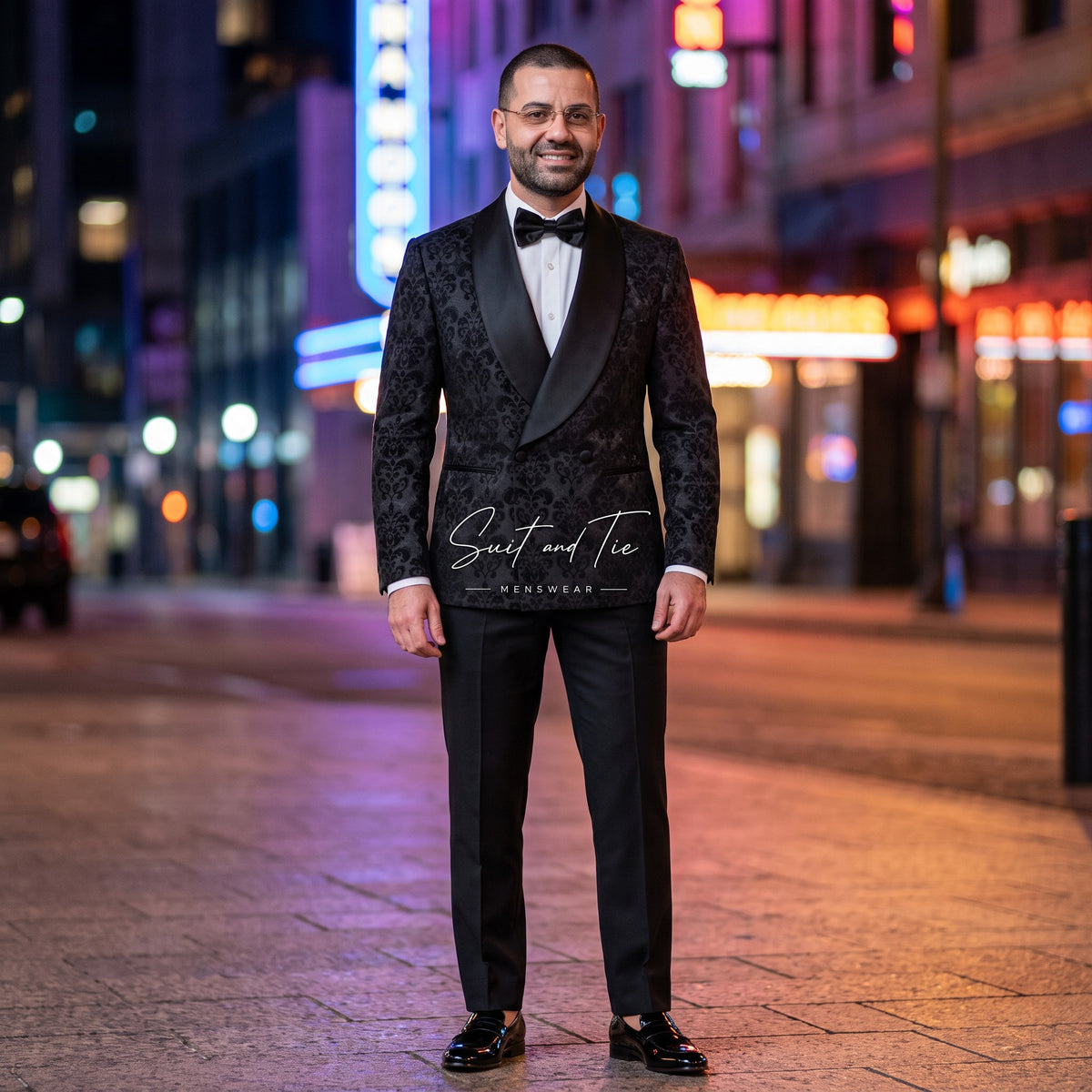 A man wearing a black patterned tuxedo with black satin lapels, a white dress shirt, and a black bow tie stands on a city street at night with blurred neon lights in the background. A watermark reading 'Saint and Tie' is visible across the lower torso.