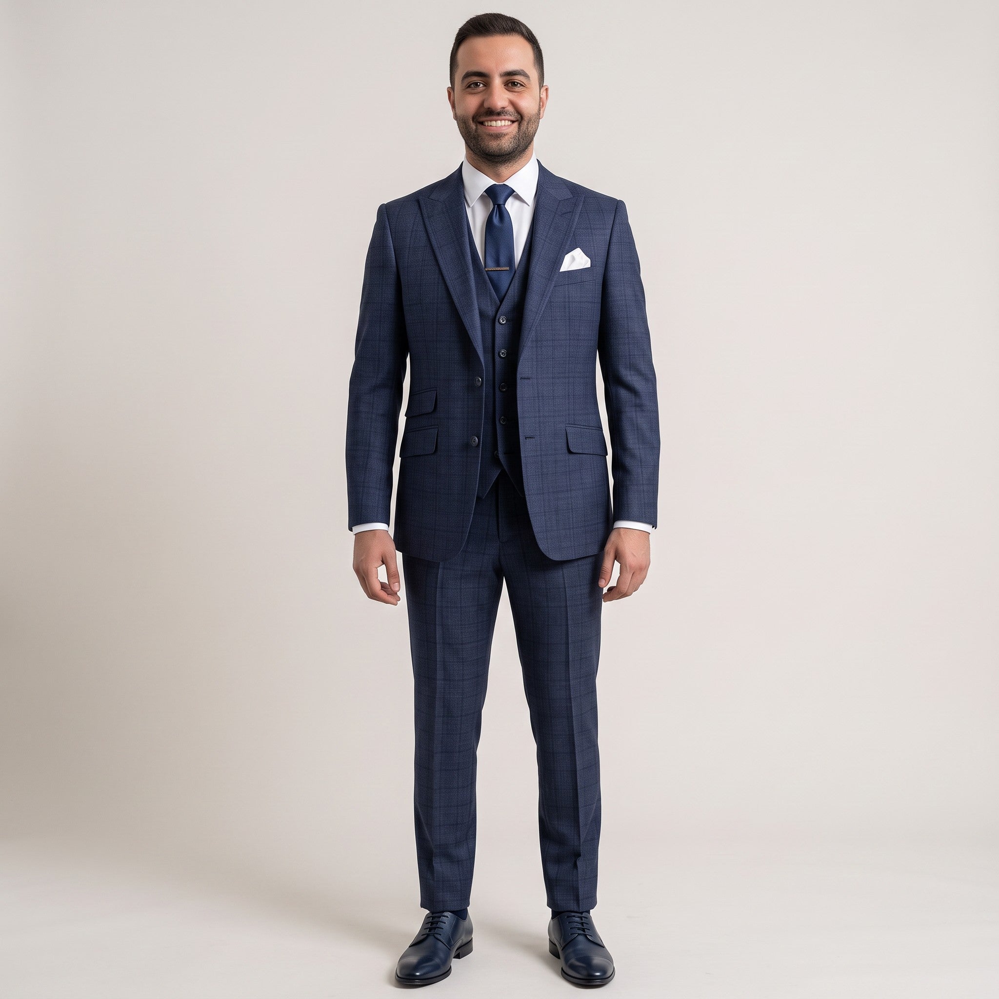 A man wearing a navy blue three-piece suit with a subtle windowpane check pattern, a matching vest, a blue patterned tie, a white pocket square, and dark dress shoes, standing against a light beige studio background.