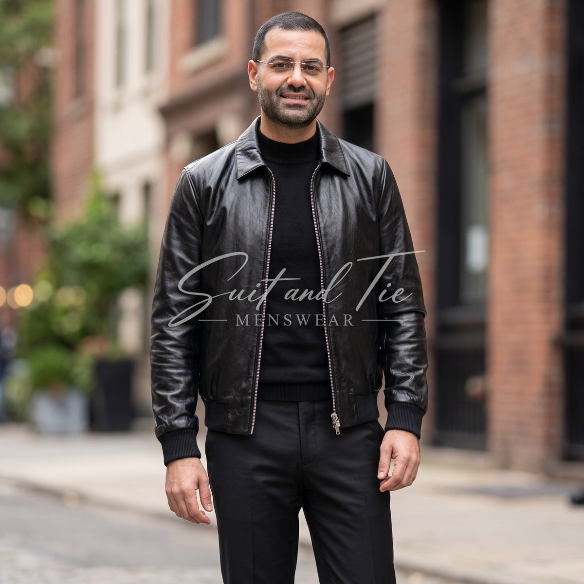 A man wearing a black leather jacket over a black turtleneck and black trousers, standing on a city street with brick buildings in the background. A 'Suit and Tie Menswear' watermark is visible across the center.