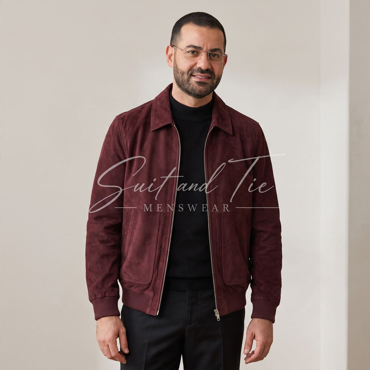 A man wearing a burgundy suede jacket over a black t-shirt and black trousers, standing against a plain beige wall. A watermark reading 'Suit and Tie MENSWEAR' is overlaid across the center.