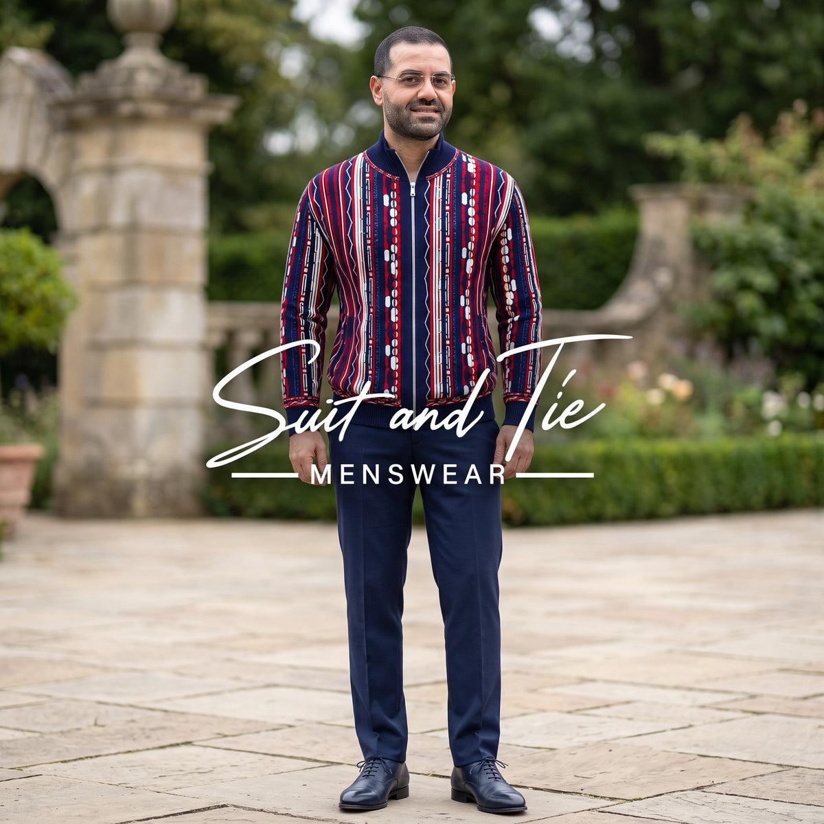 A man wearing a patterned red, navy, and white striped shirt with navy trousers stands in a garden courtyard. A large 'Suit and Tie MENSWEAR' logo overlay is centered on the image.