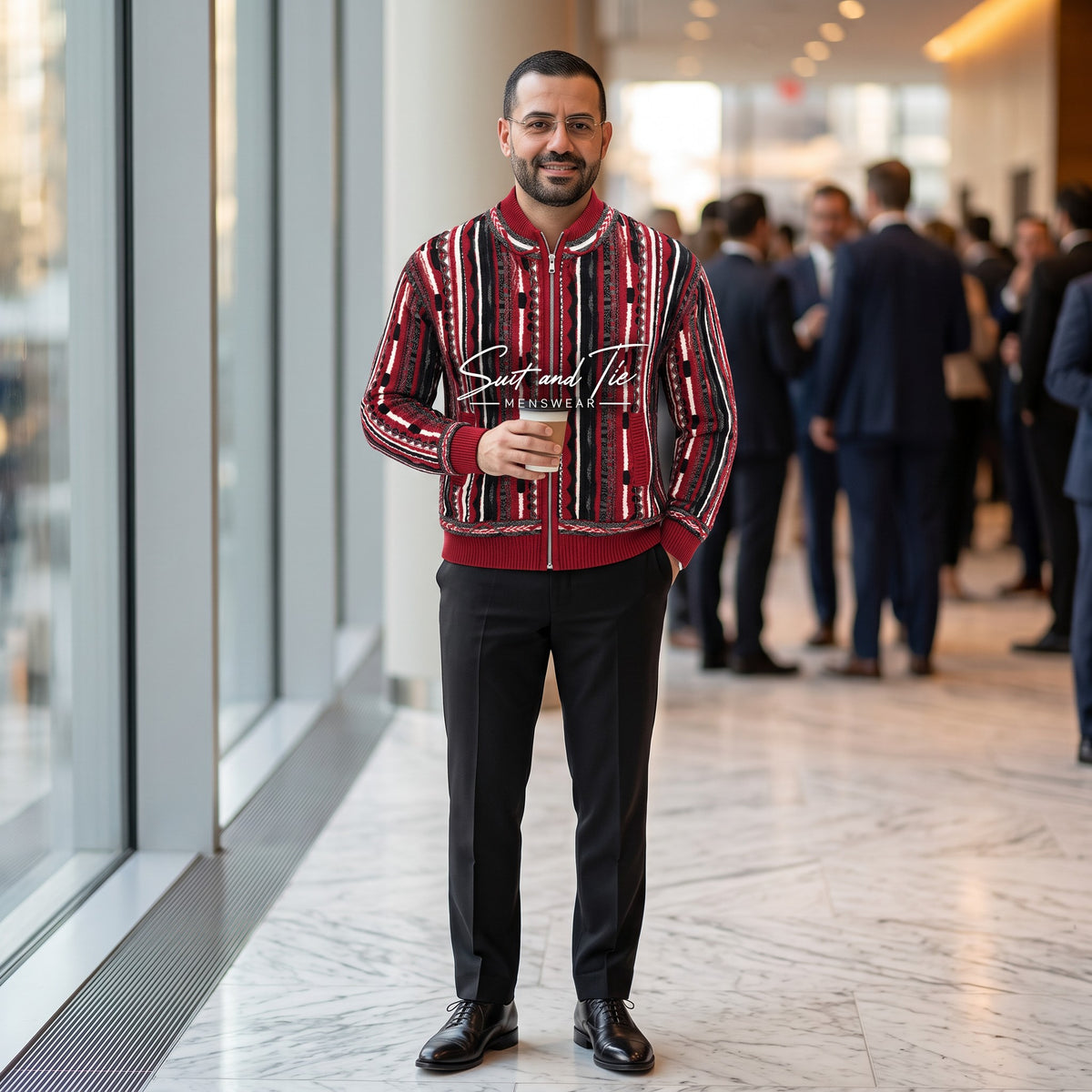 A man wearing a red and black patterned bomber jacket with a high collar, standing in a modern office lobby. He is holding a coffee cup and wearing black trousers and dress shoes.