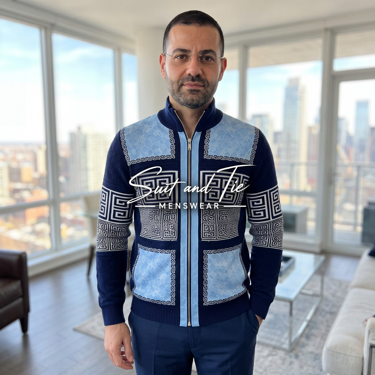 A man wearing a blue and navy patterned zip-up sweater with Greek key motifs, standing in a modern high-rise apartment with a city skyline view. A 'Suit and Tie MENSWEAR' watermark is visible across the center.