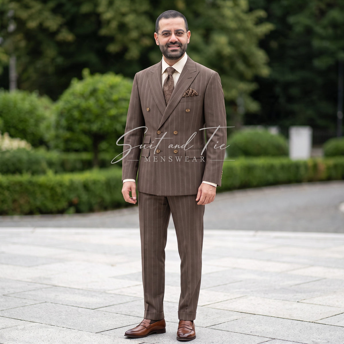 A man wearing a brown double-breasted pinstripe suit with a matching tie and pocket square, standing outdoors on a paved walkway with green trees in the background. A watermark for 'Giacco and Tie Menswear' is visible across the center.