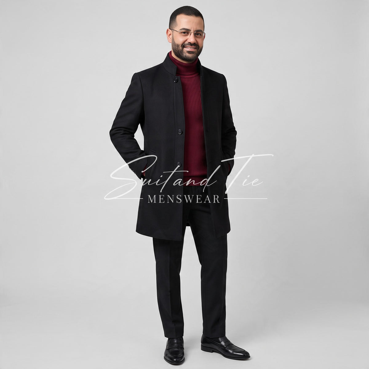 A man modeling a black men's overcoat with a mandarin collar, worn over a dark red turtleneck sweater, black trousers, and black dress shoes, against a light gray studio background. A 'Suitand Tie Menswear' watermark is visible across the center.