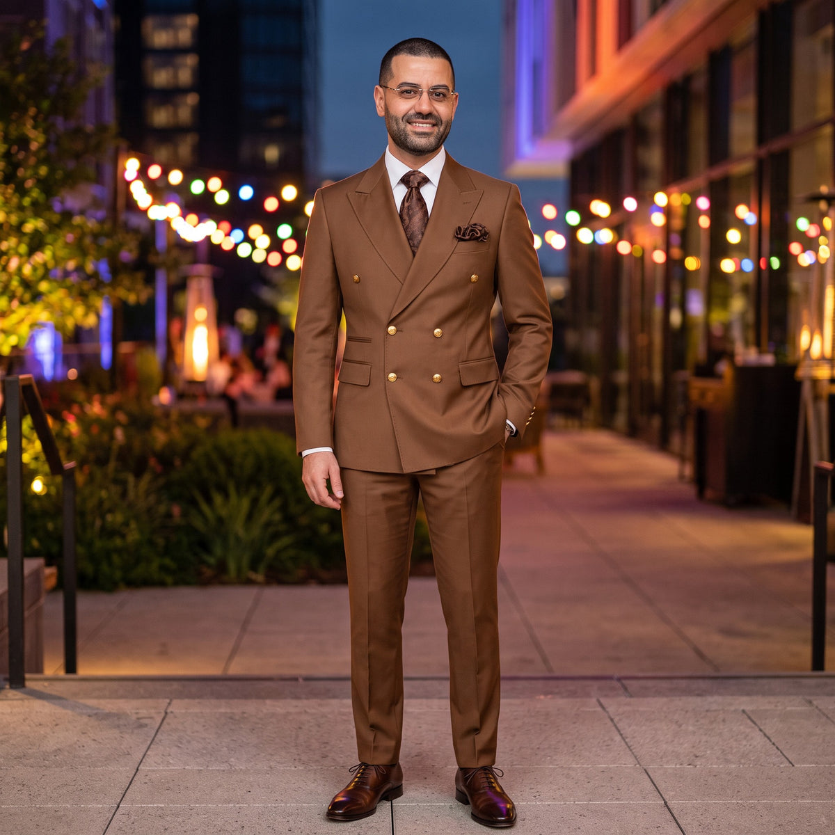 A man wearing a brown double-breasted suit with gold buttons, a white shirt, and a brown patterned tie, standing in a city outdoor setting at night with blurred string lights in the background.