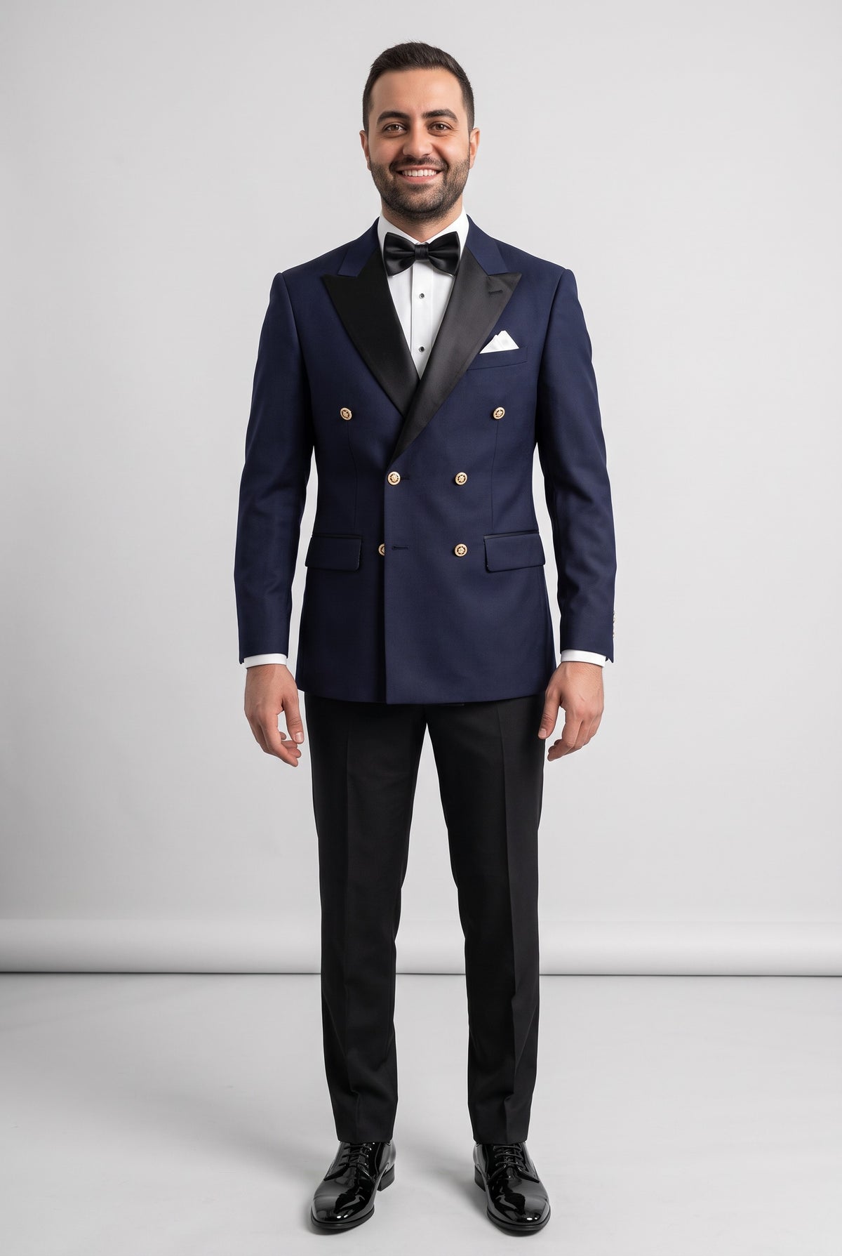 A man modeling a navy blue double-breasted tuxedo jacket with black satin lapels, gold buttons, black trousers, a white dress shirt, a black bow tie, and a white pocket square, set against a plain light gray studio background.
