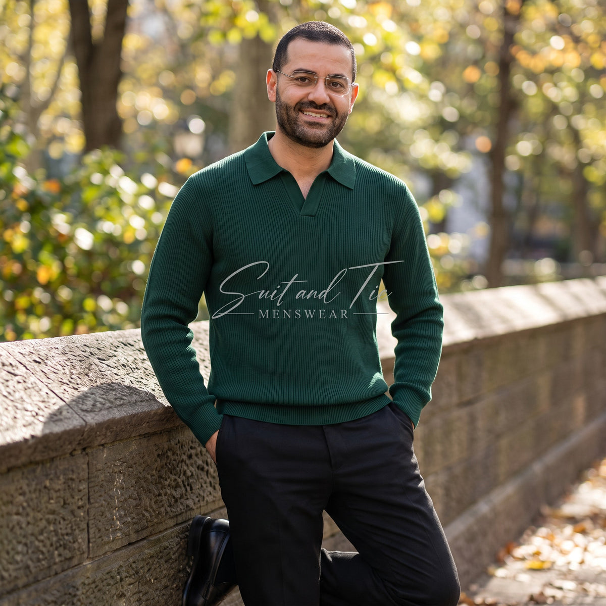 A man wearing a dark green ribbed polo sweater and black trousers, leaning against a stone wall in an outdoor park setting with autumn trees in the background.