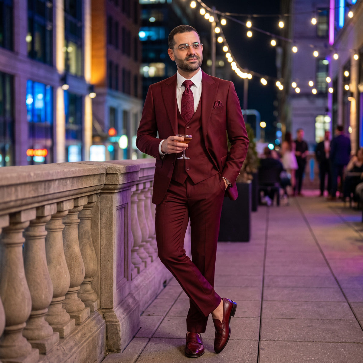 A man wearing a three-piece burgundy suit, white dress shirt, and matching burgundy tie stands on a city sidewalk at night, holding a glass. The background features a stone balustrade, warm string lights, and blurred city buildings.