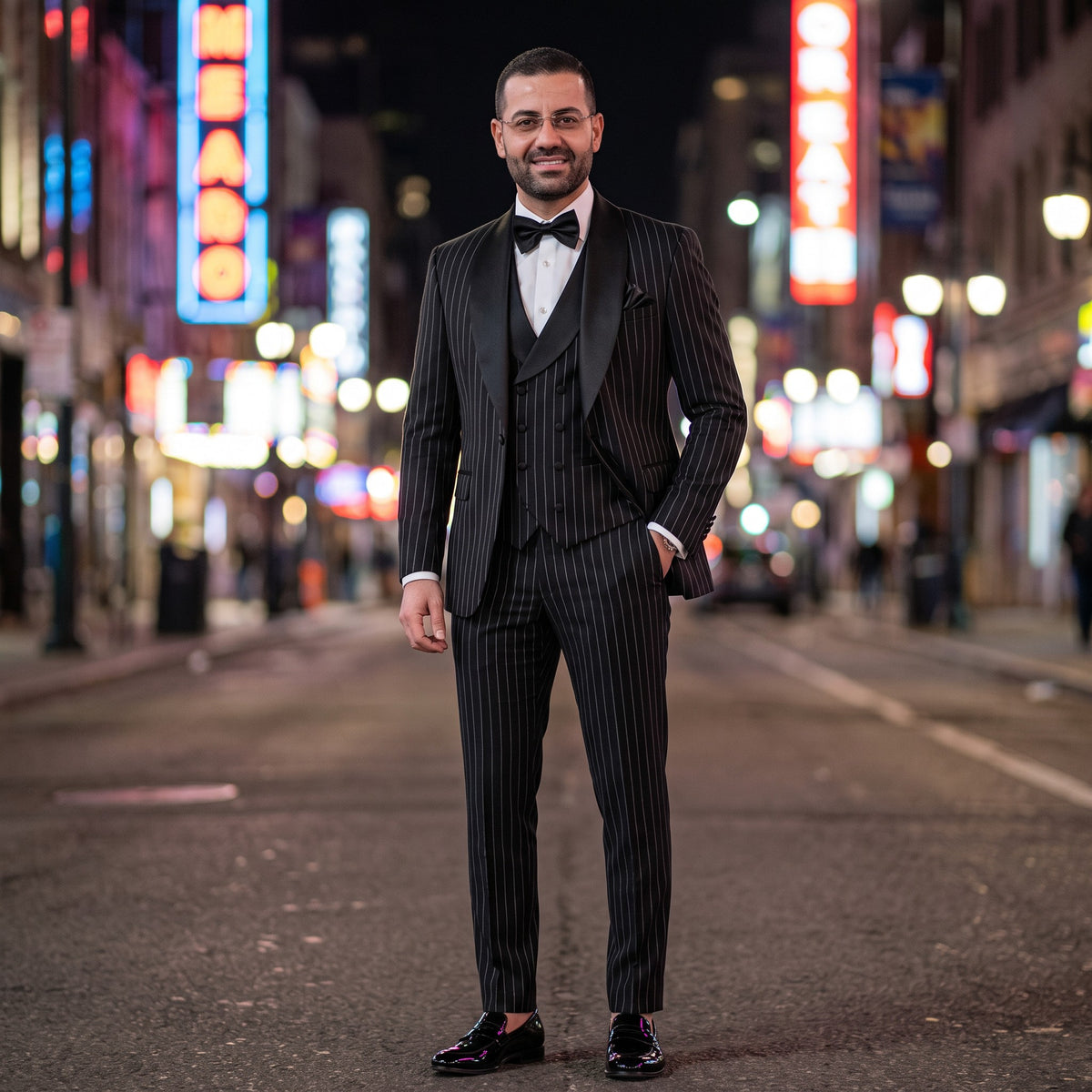 A man wearing a black pinstripe three-piece tuxedo with a white shirt, black bow tie, and black dress shoes stands on a city street at night with blurred neon signs in the background.