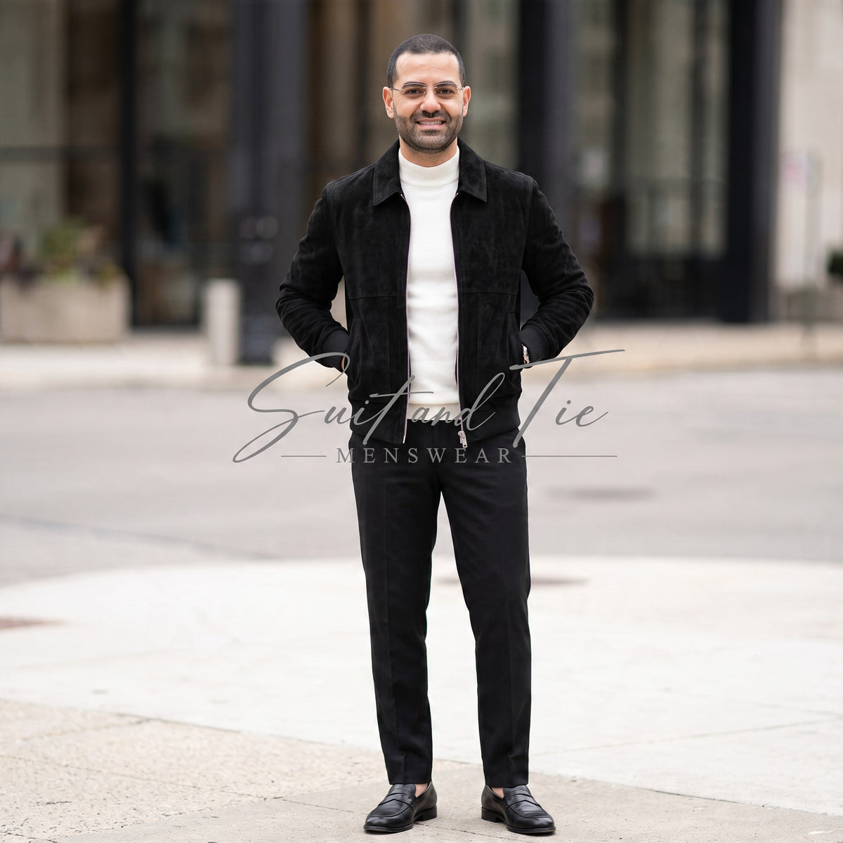 A man wearing a black corduroy jacket over a white turtleneck sweater, black trousers, and black loafers, standing outdoors on a city sidewalk. A 'Suit and Tie Menswear' watermark is visible across the center.