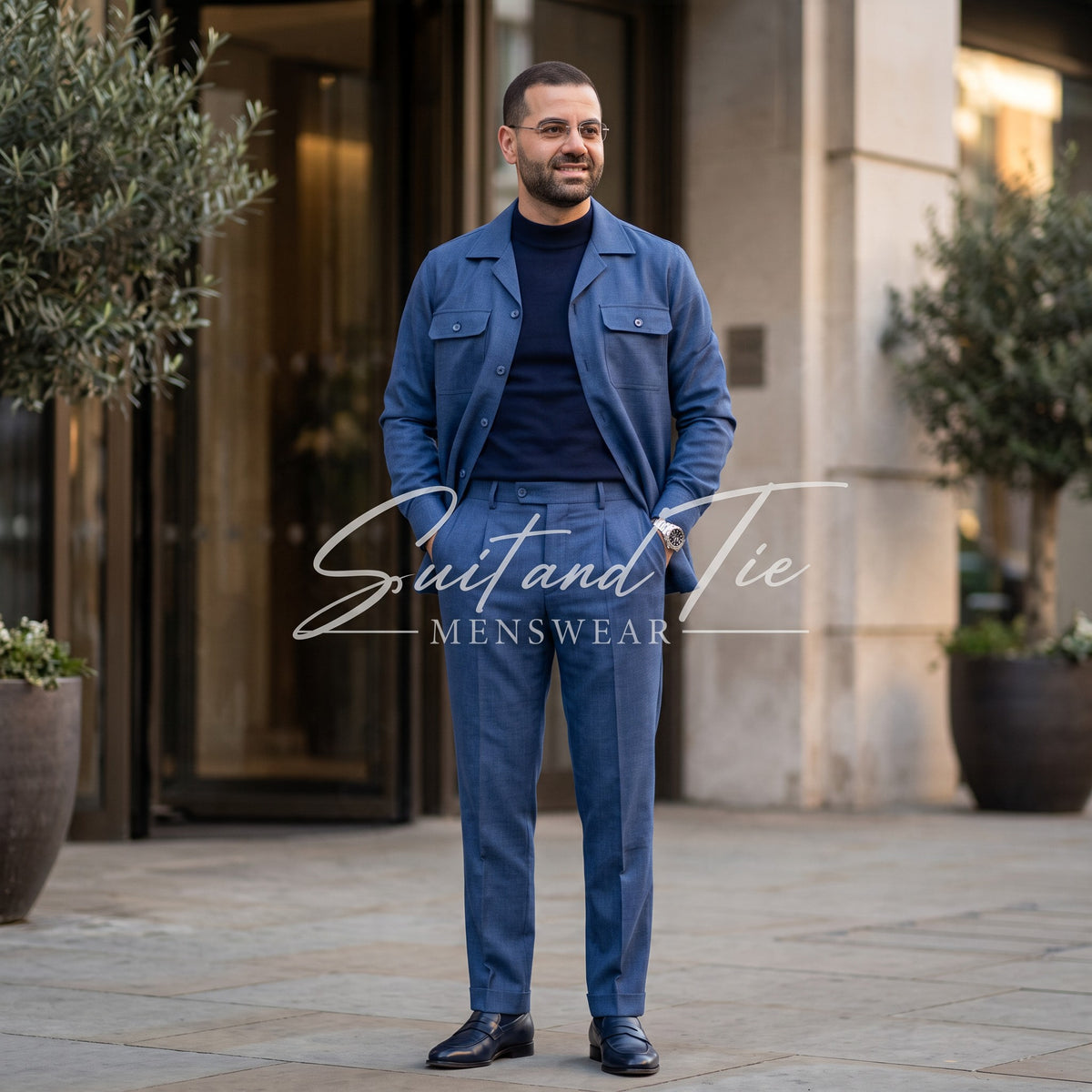 A man wearing a blue two-piece suit jacket and matching trousers over a dark navy turtleneck, standing outdoors in front of a building entrance. A white script watermark reading 'Suit and Tie MENSWEAR' is overlaid across the center.