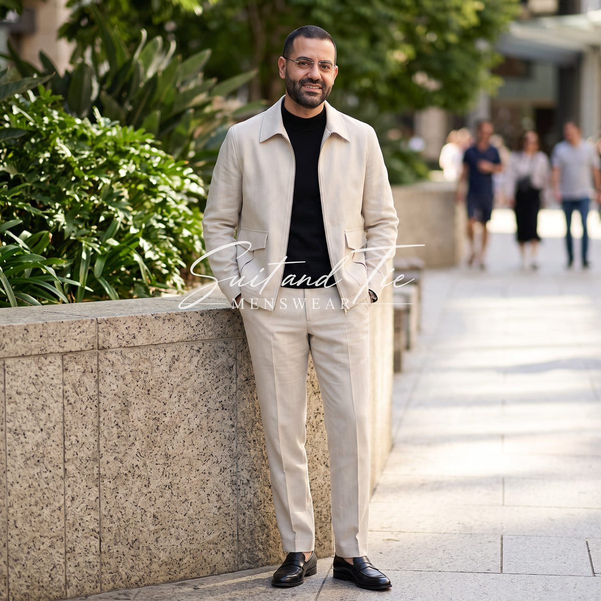 A man wearing a light beige two-piece suit with a black t-shirt and black loafers, standing outdoors on a paved walkway next to a stone planter.