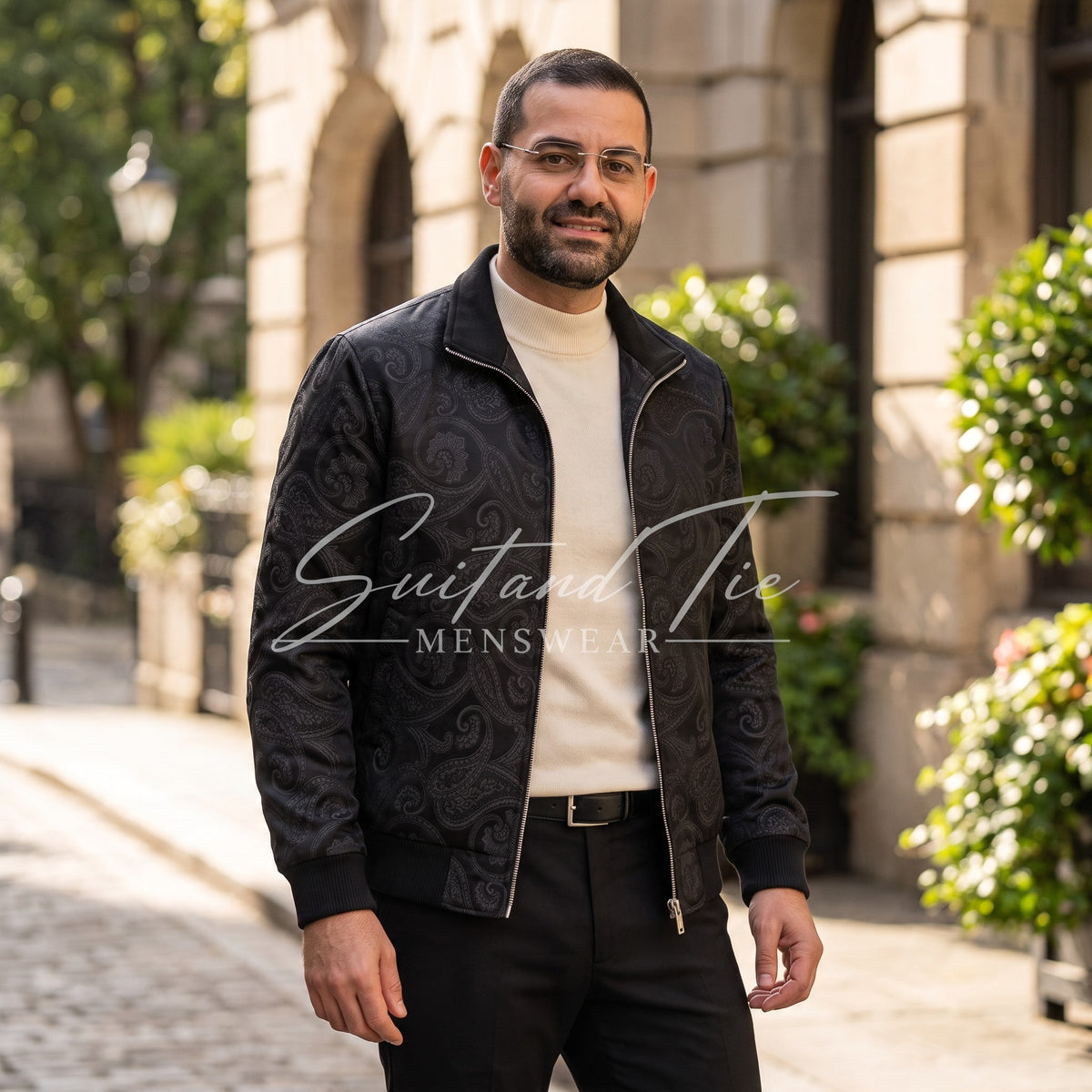 A man wearing a black jacquard patterned bomber jacket over a cream turtleneck sweater and black trousers, standing on a cobblestone street in front of a stone building. A watermark for 'Suit and Tie MENSWEAR' is overlaid across the center.