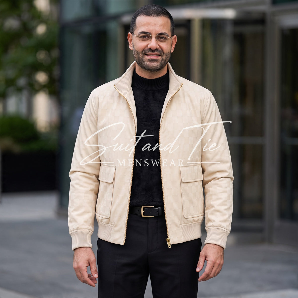 A man wearing a light beige suede jacket over a black turtleneck and black trousers, standing outdoors in front of a modern glass building. A watermark reading 'Suit and Tie Menswear' is overlaid across the center.