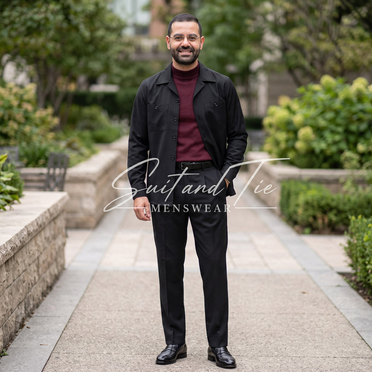 A man wearing a black overshirt, burgundy turtleneck, black trousers, and black dress shoes, standing on a paved walkway in a garden setting. A watermark for 'Suit and Tie Menswear' is overlaid across the center.