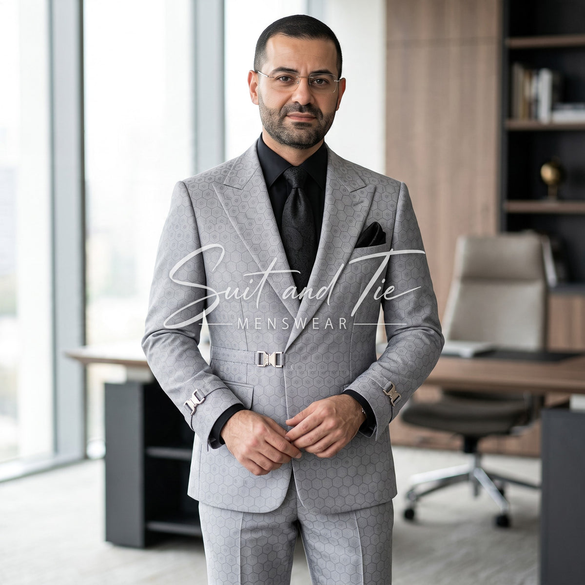 A man wearing a light gray patterned suit, black shirt, and black tie stands in a modern office setting. A large white script watermark reading 'Suit and Tie MENSWEAR' is overlaid across the center.