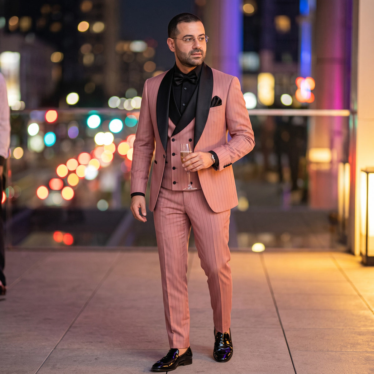 A man wearing a pink pinstripe three-piece tuxedo with black satin lapels, a black bow tie, and black dress shoes, standing on a balcony at night with a blurred city lights background.