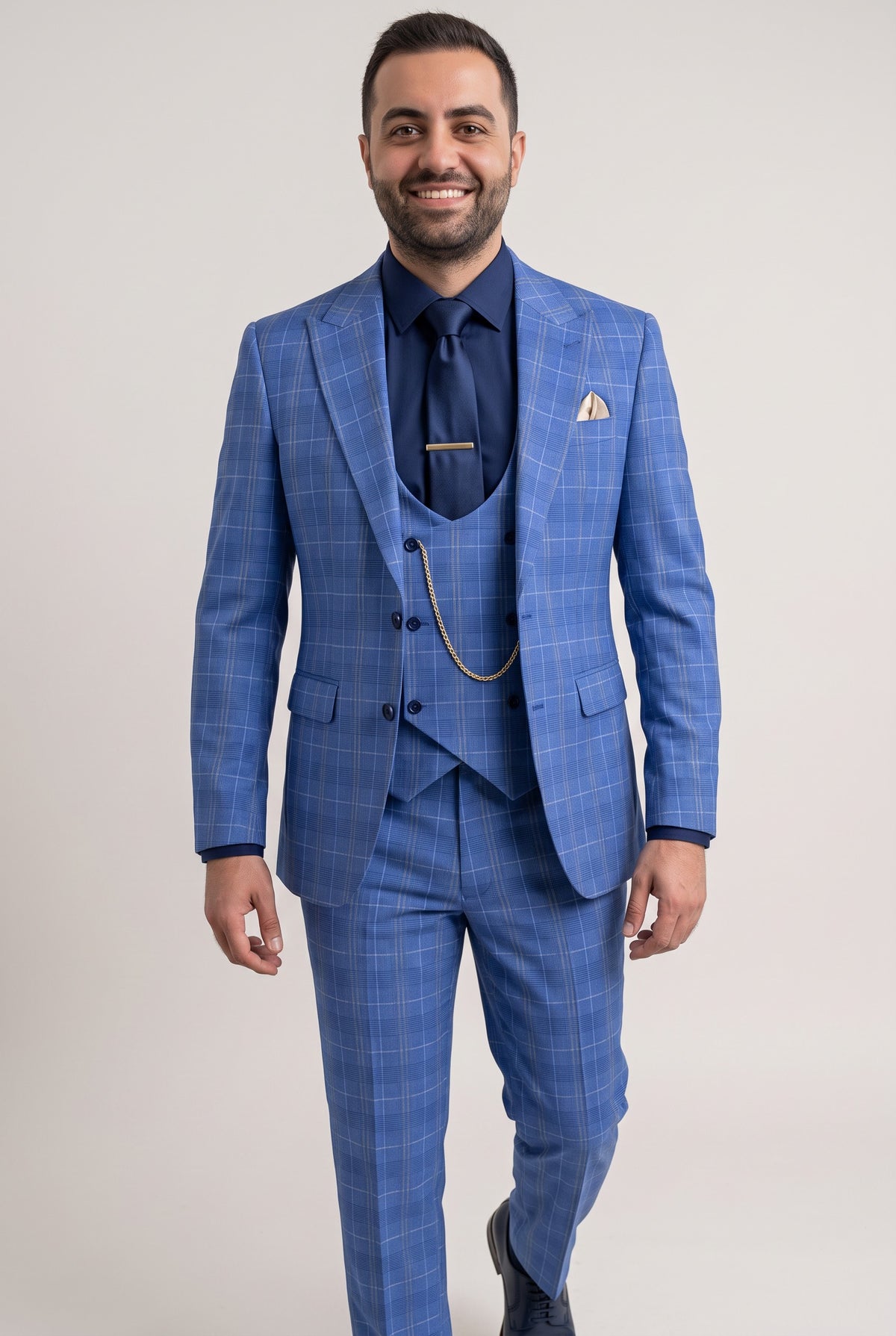 A man wearing a blue three-piece suit with a subtle windowpane plaid pattern, paired with a navy blue shirt, tie, and pocket square, standing against a light gray studio background.