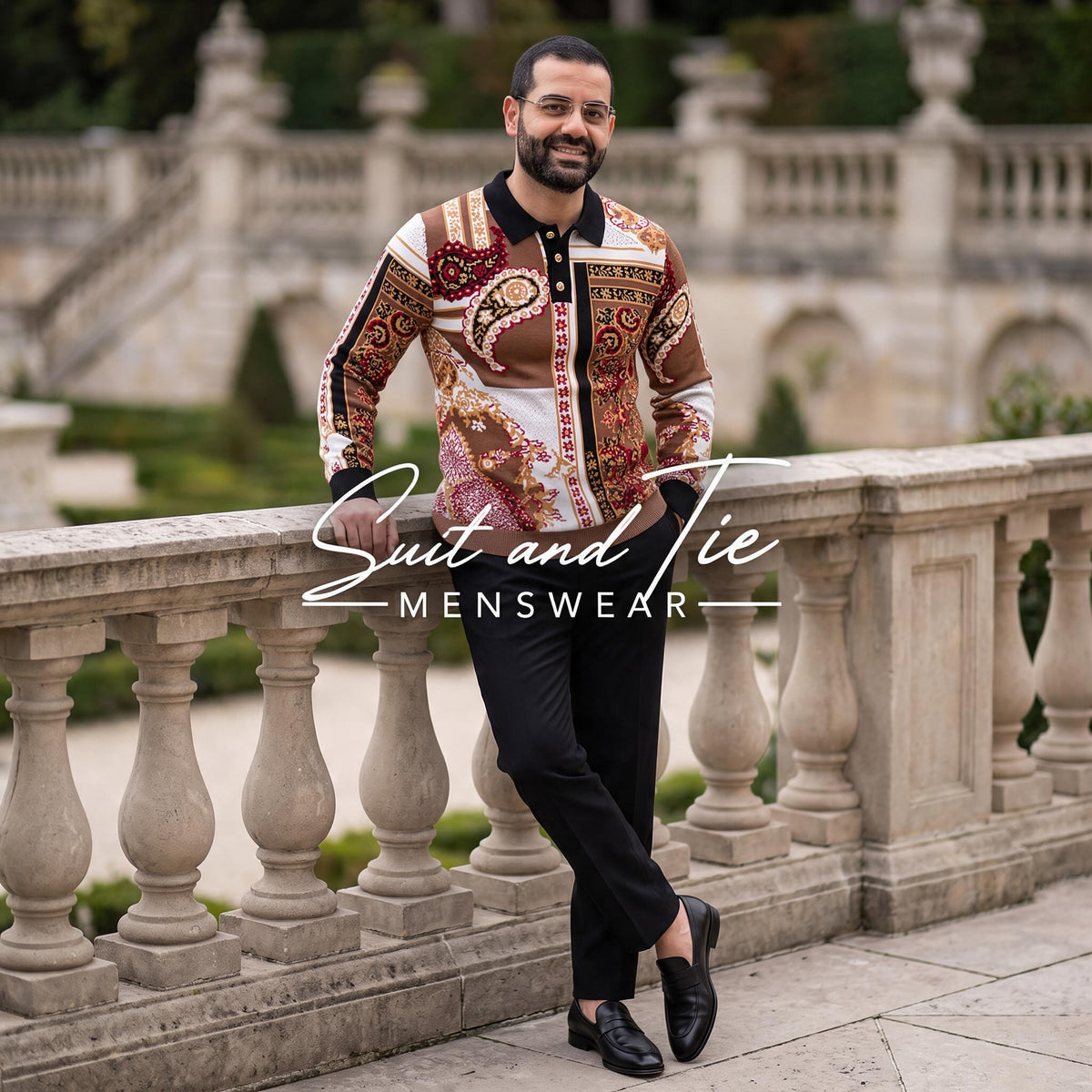 A man wearing a patterned brown and beige Suit and Tie Menswear shirt, black trousers, and black loafers, leaning against a stone balustrade in an outdoor garden setting.