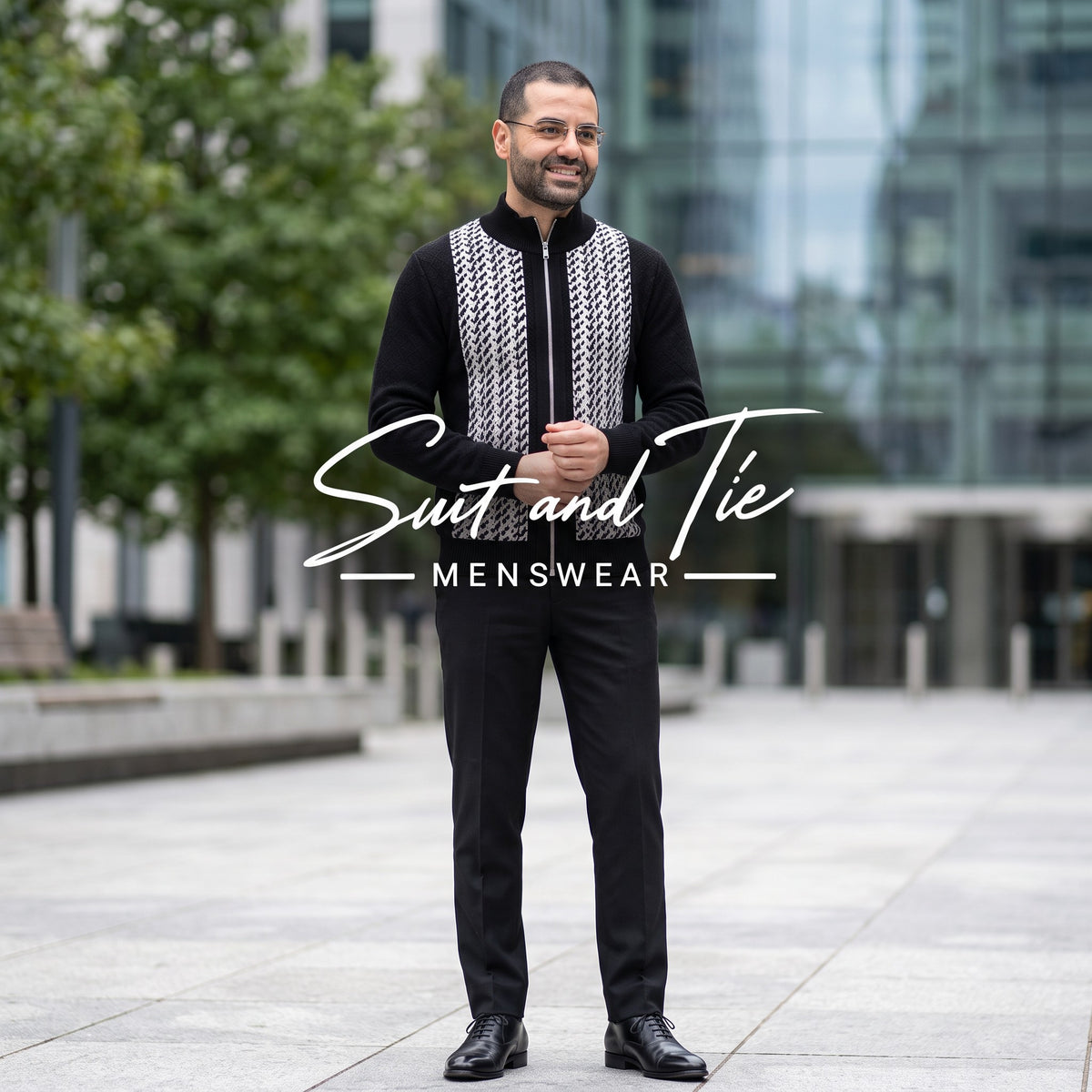 A man wearing a black men's suit with a black and white geometric patterned vest and black trousers, standing outdoors in a modern city setting. A watermark overlay reads 'Suit and Tie MENSWEAR'.