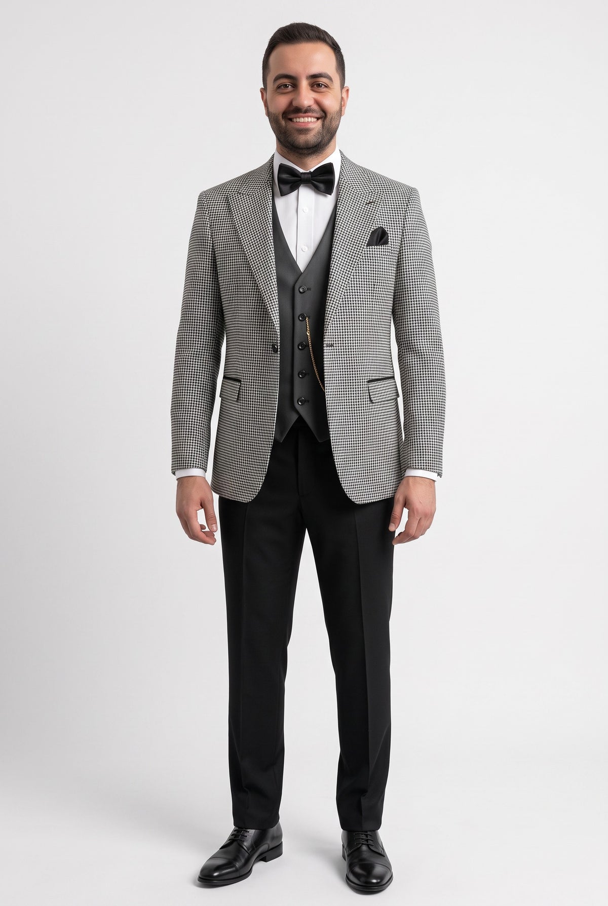 A man modeling a grey and white micro-check suit jacket with a matching grey vest, black trousers, a black bow tie, and a white dress shirt against a light grey studio background.