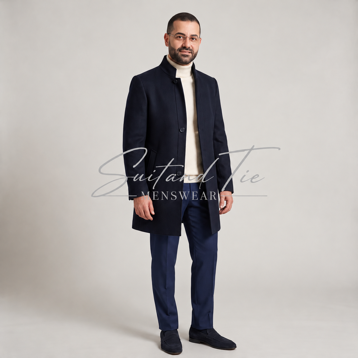 A man modeling a dark navy blue wool-blend coat over a cream turtleneck sweater, navy trousers, and dark loafers, standing against a light gray studio background. A 'Suit and Tie Menswear' watermark is visible across the center.