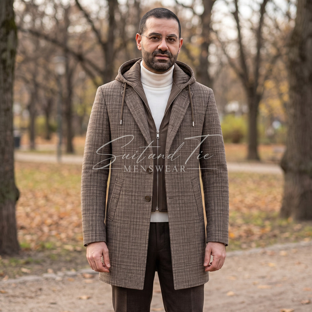A man wearing a brown and beige plaid hooded coat over a white turtleneck sweater and dark trousers, standing in a park with blurred trees in the background. A watermark reading 'SuitandTie Menswear' is visible across the center.