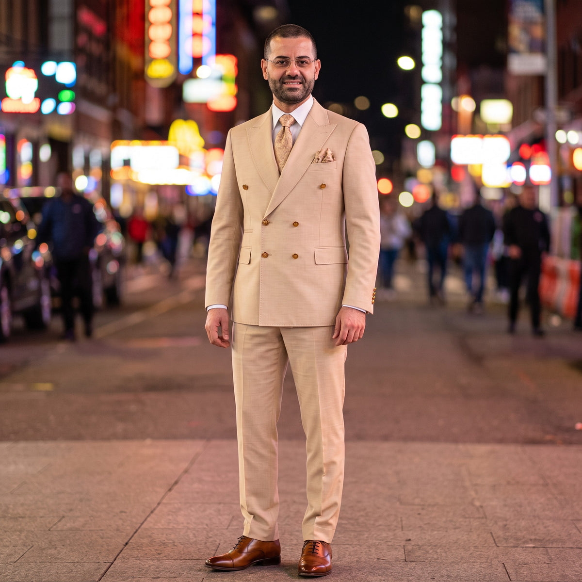 A man wearing a beige double-breasted suit with brown dress shoes stands on a city street at night with blurred neon lights in the background.
